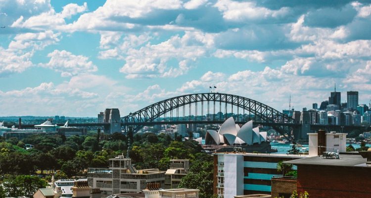Sydney skyline featuring the Harbour Bridge and the Opera House against a bright blue sky.