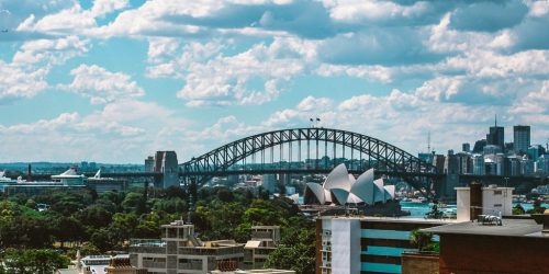 Sydney skyline featuring the Harbour Bridge and the Opera House against a bright blue sky.